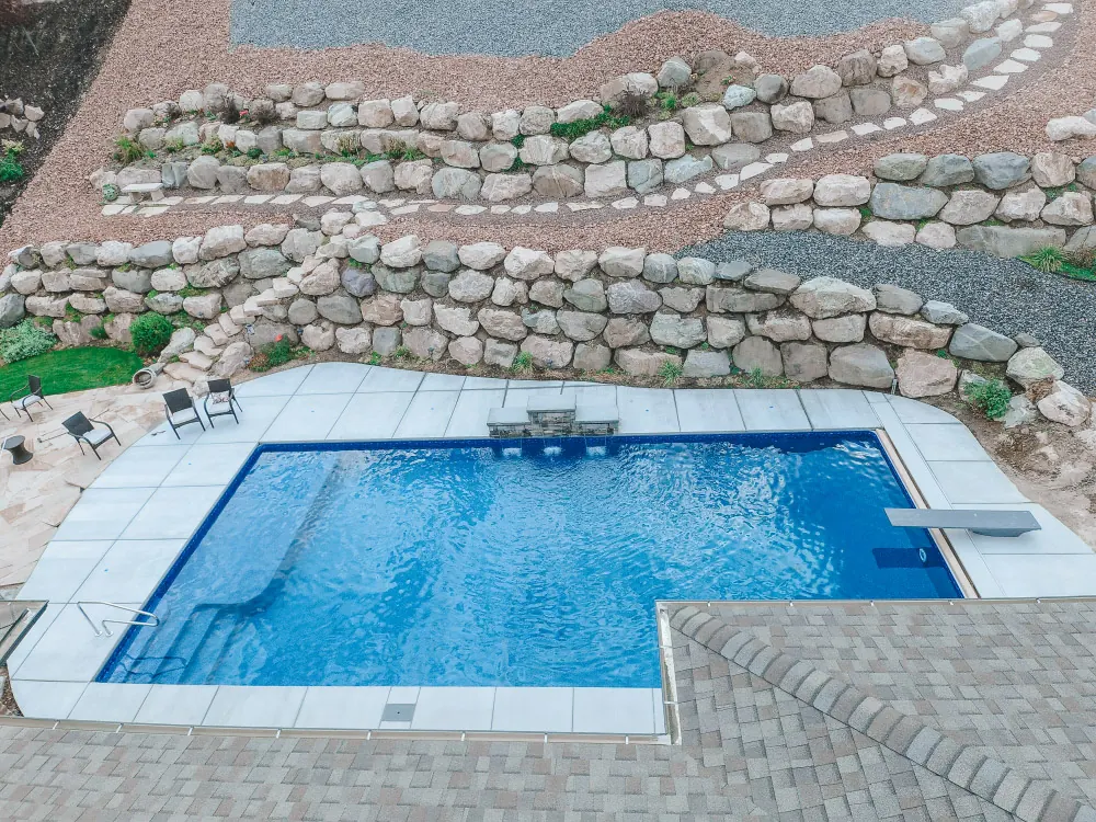 Layton Utah Aerial View Vinyl Pool with Boulder Retaining Wall - Barr Pool Builders Aerial drone view of a custom rectangle vinyl swimming pool with diving board, stone patio and terraced boulder retaining wall in Layton, Utah by Barr Pool Builders