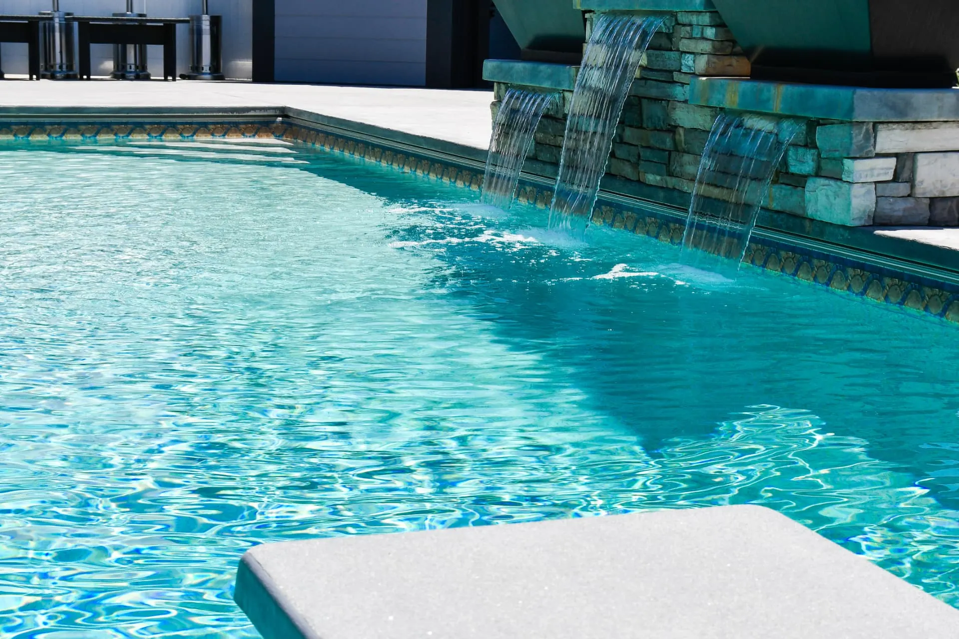 Layton Utah Vinyl Pool with Stone Waterfall Close-Up - Barr Pool Builders Close-up of stone waterfall feature cascading into a custom vinyl swimming pool with mosaic tile trim and diving board in Layton, Utah by Barr Pool Builders