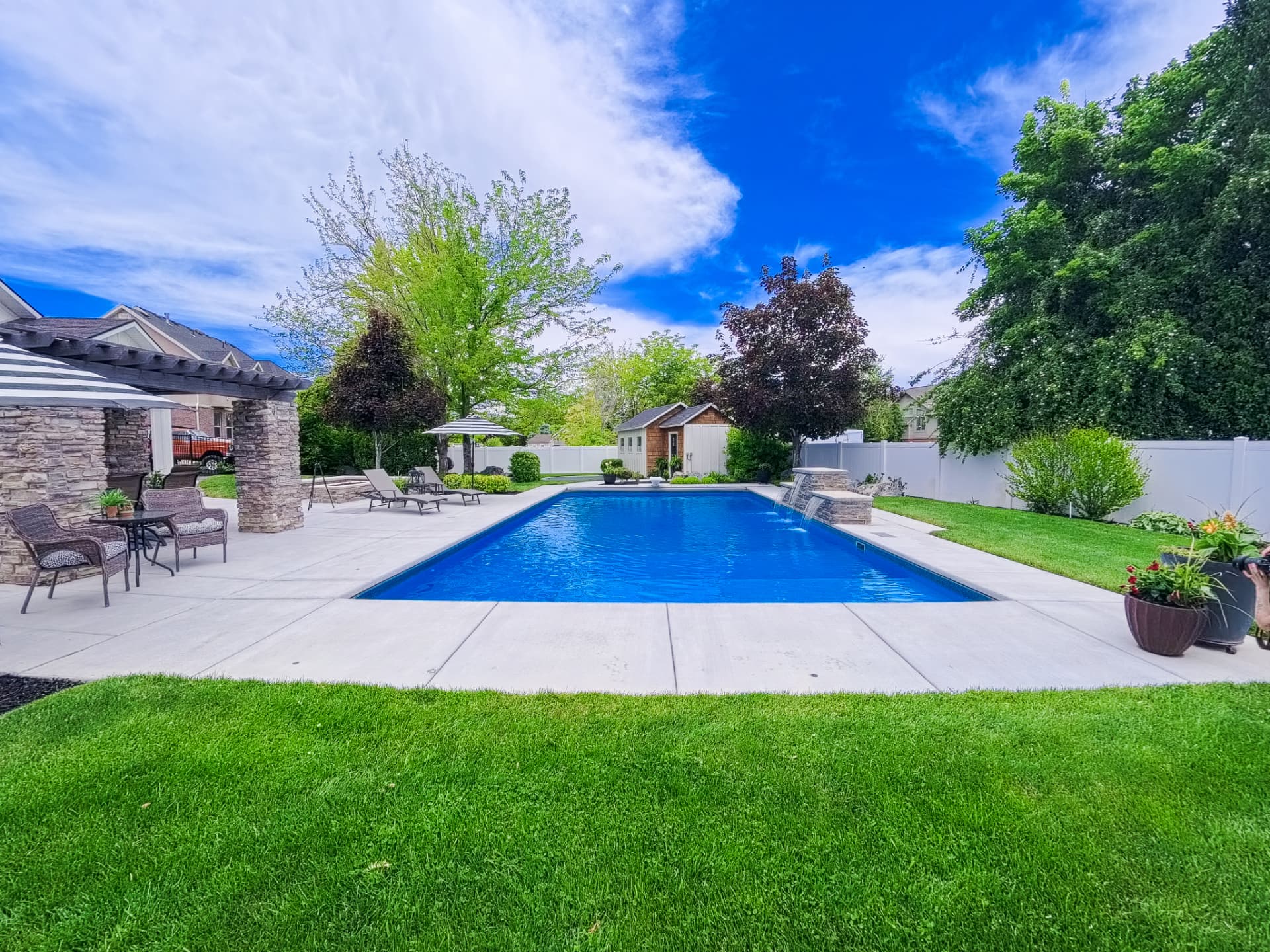 Syracuse Utah Rectangle Vinyl Pool with Pergola and Water Features - Barr Pool Builders Rectangle vinyl inground swimming pool with stone pergola, striped umbrella lounge and cascading water features in a Syracuse, Utah backyard by Barr Pool Builders